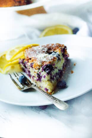 slice of blueberry yogurt cake sitting on a white plate with a fork and garnished with lemon slices