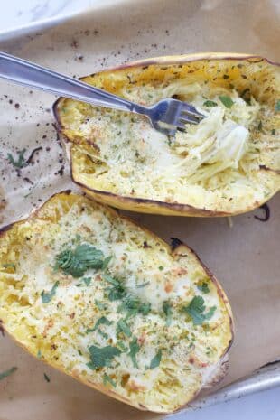 two spaghetti squash halves with melted cheese and herbs and a fork with twisted spaghetti squash